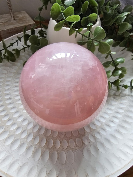 Large polished Rose Quartz sphere crystal displaying soft pink hue and translucent quality, photographed on white textured surface with green eucalyptus leaves, perfect for meditation and heart chakra healing